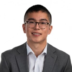 Jevons Liu is Baltimore City Public Schools' Teacher of the year. He is wearing a suit and glasses while smiling at the camera.